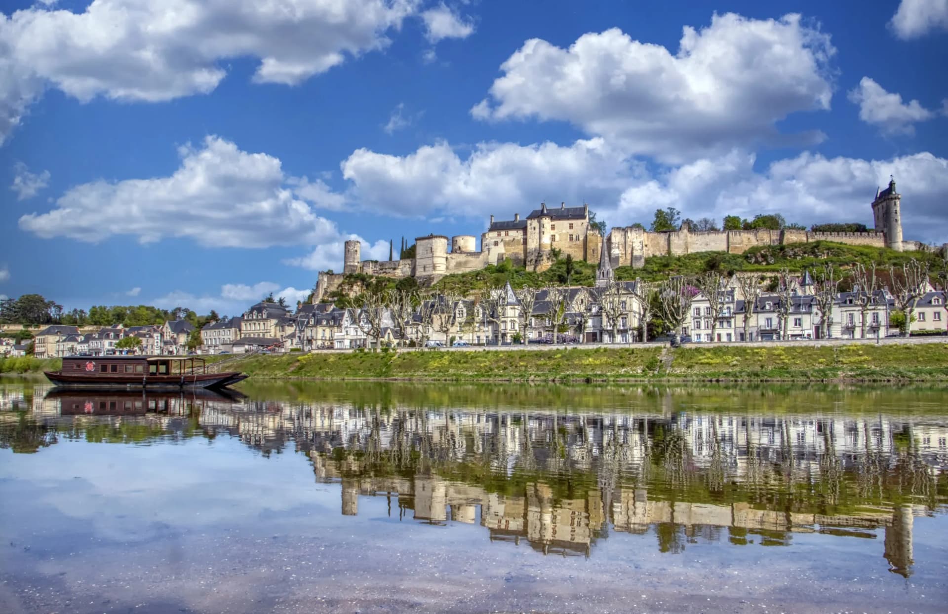 Panoramic view of Chinon city with royal castle above houses, reflected in the river with a boat.