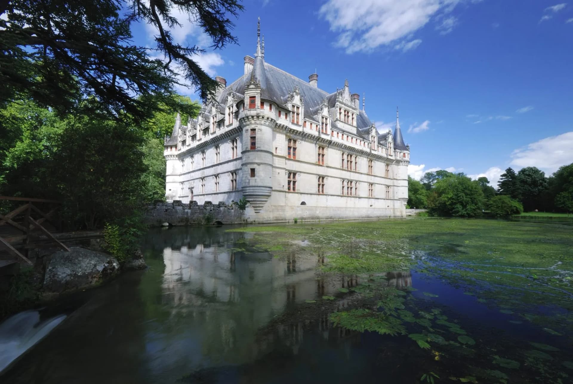 Renaissance-style chateau with turrets reflected in moat water under a blue, cloudy sky.