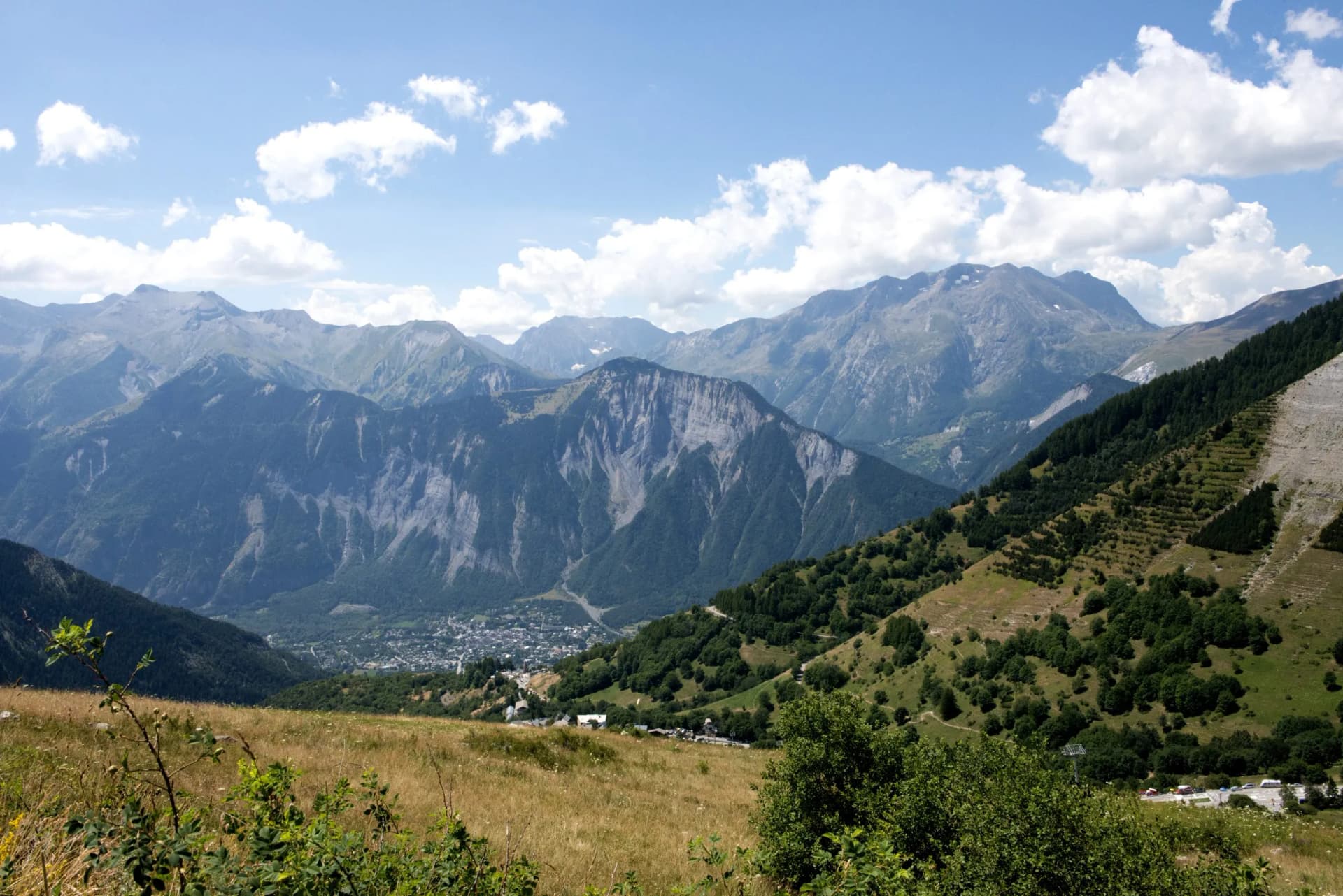 View from Les Deux Alpes, France, showing a valley town nestled among steep, forested mountains under a blue summer sky.