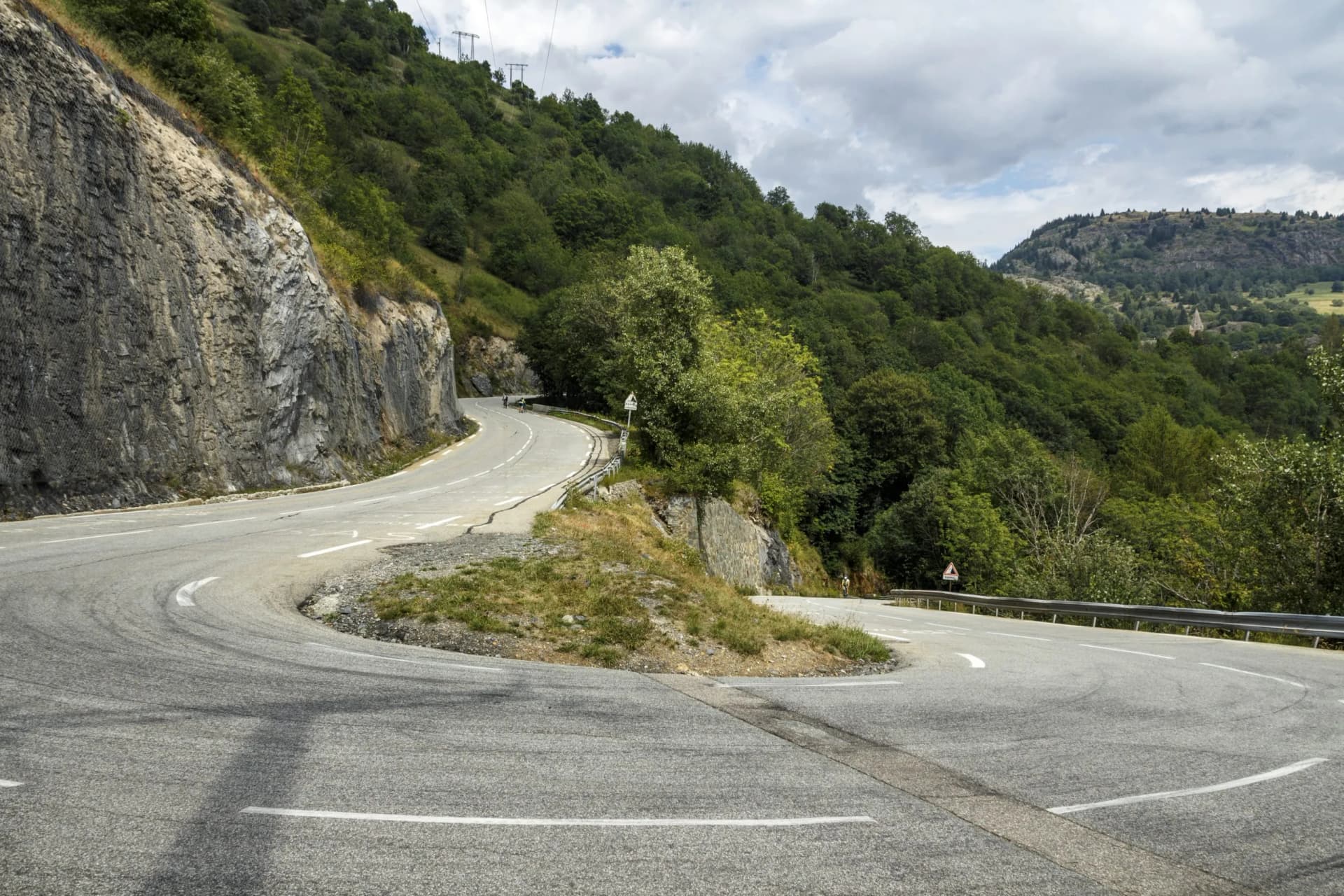 Cyclists ascending a sharp switchback road on a steep, forested mountain slope in L'Alpe d'Huez, France.