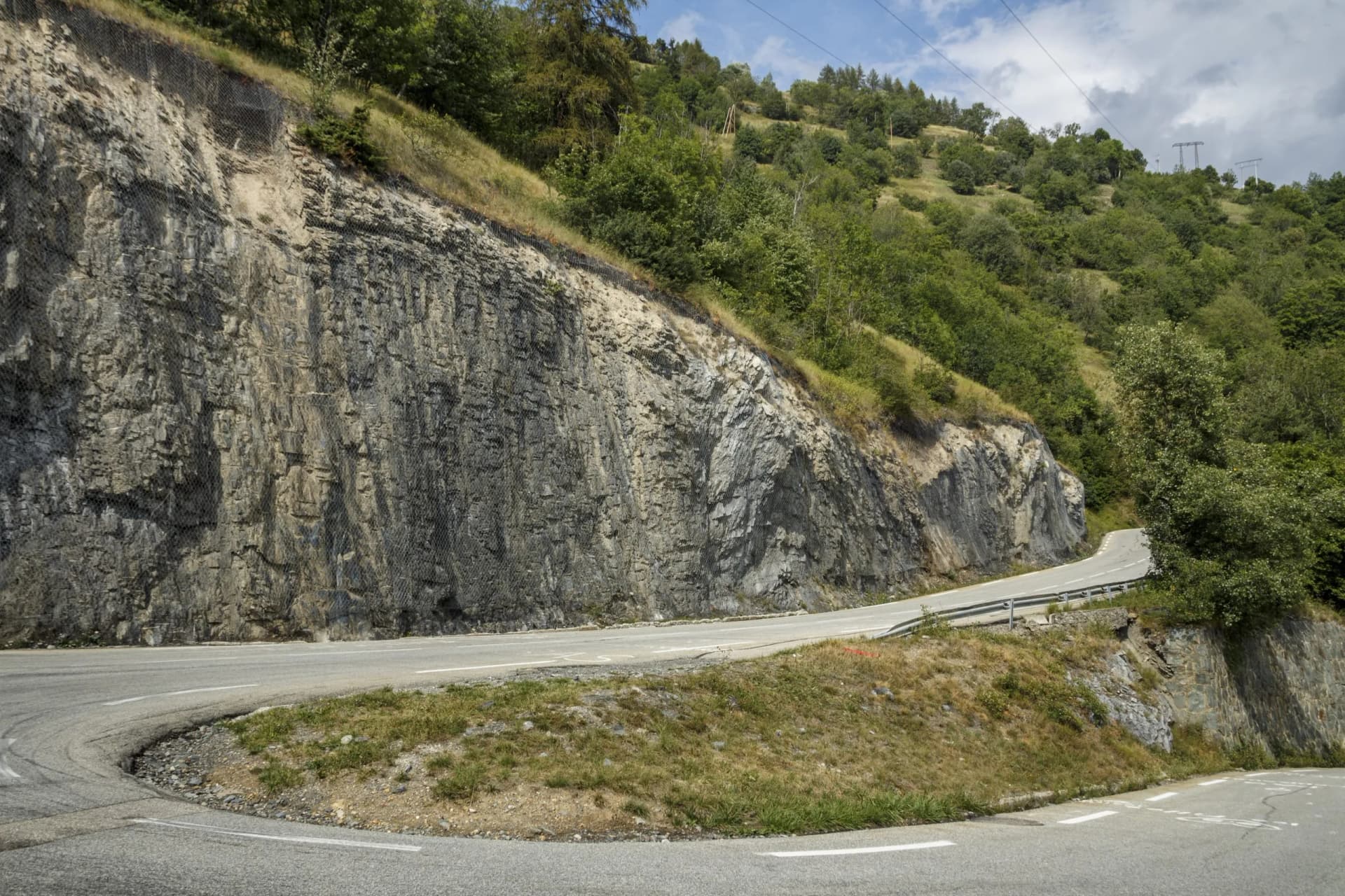 Winding mountain road ascending past a rock face with protective netting near Alpe d'Huez, France.