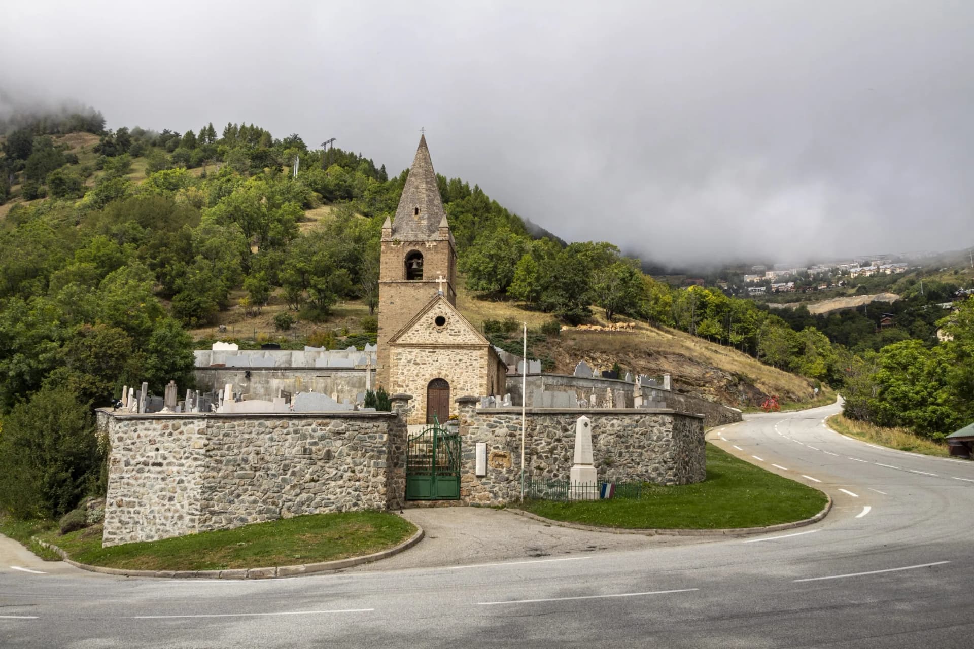 Stone church and cemetery by winding road below foggy mountains near Alpe d'Huez.