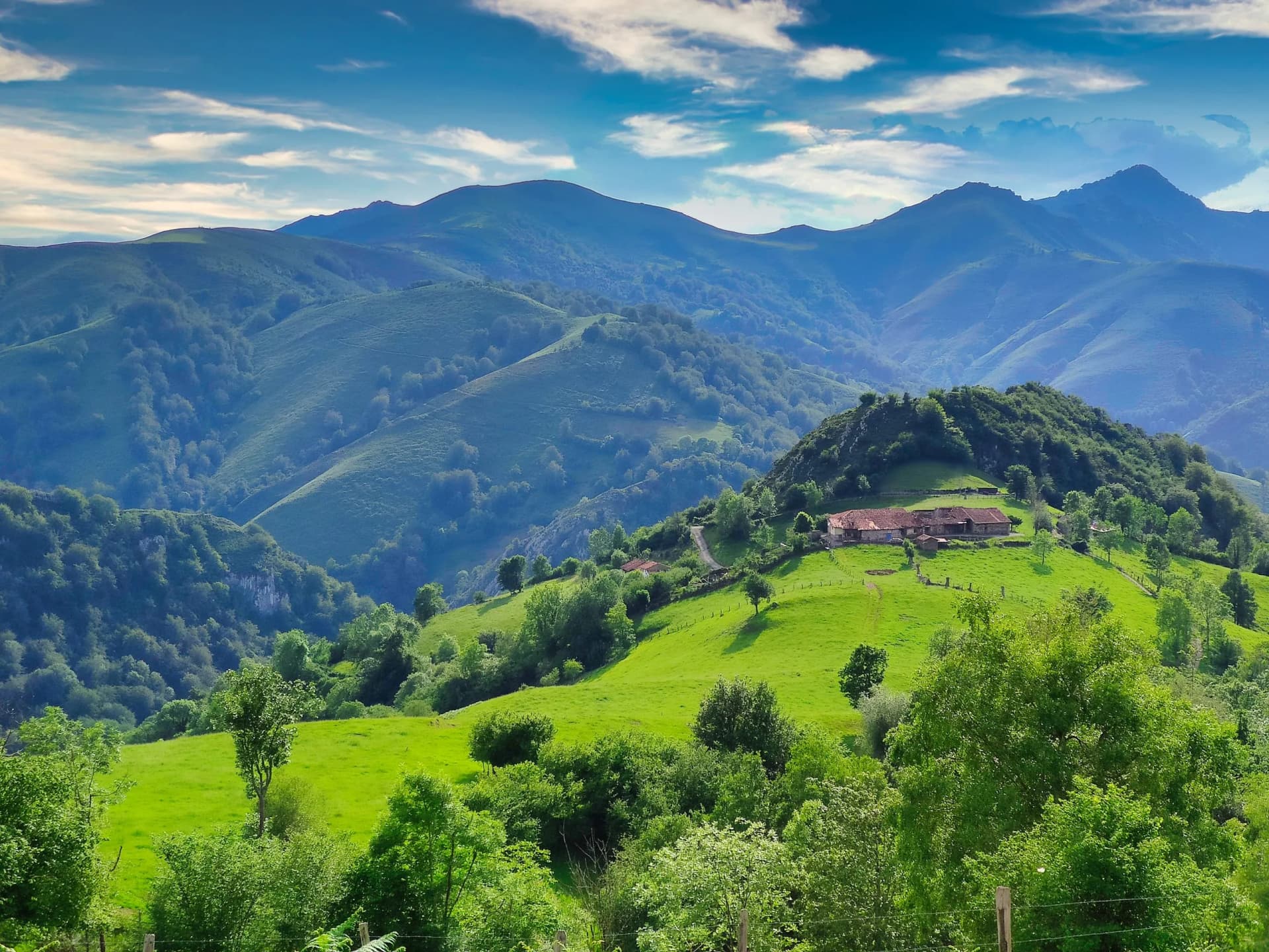 Green rolling hills and farm buildings under a blue sky in Les Tables Village, Sobrescobio Municipality, Asturias, Spain.