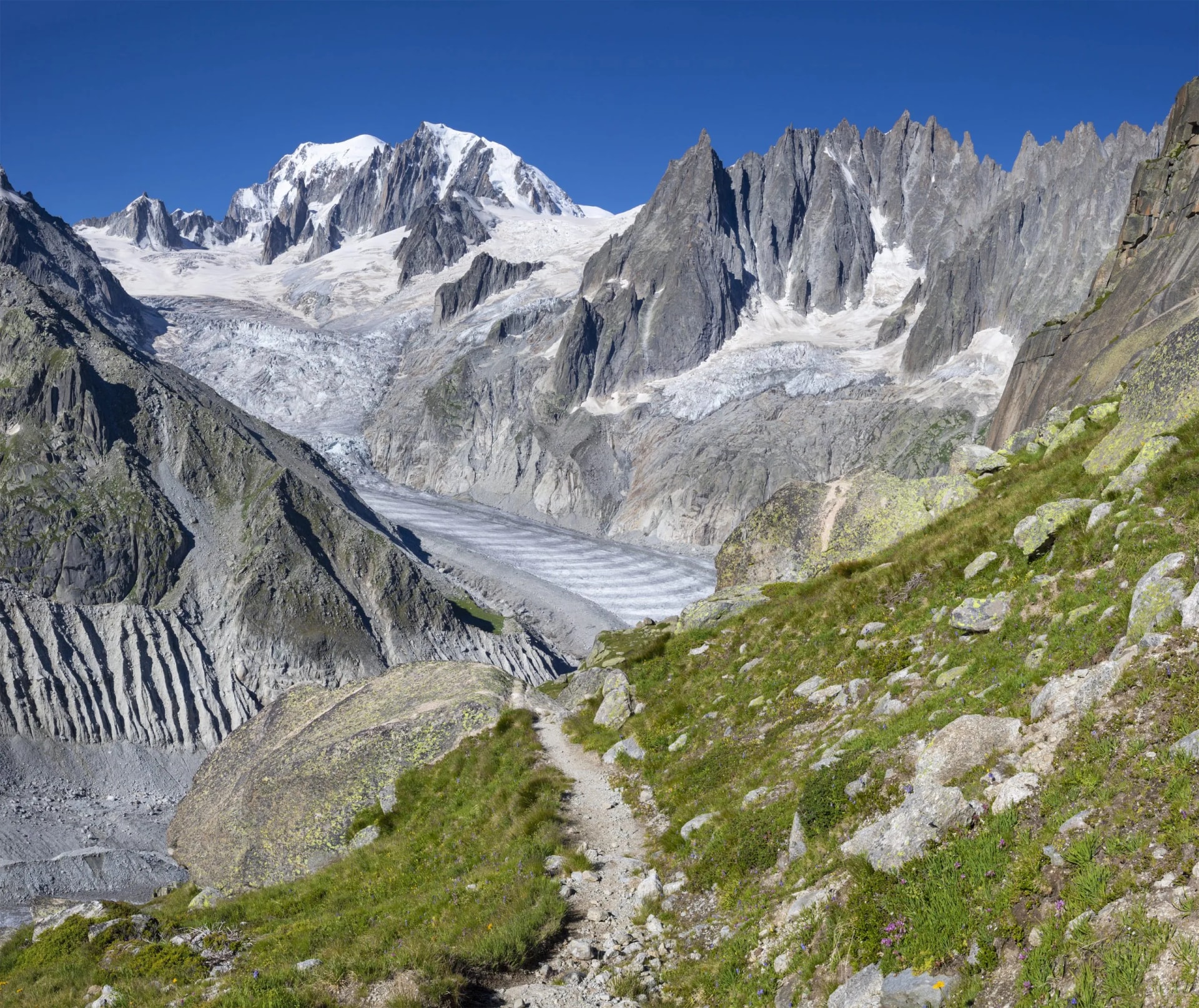 Hiking trail above glacier leading to snow-capped Mont Blanc Massif and Aiguilles towers in the Savoy Alps.