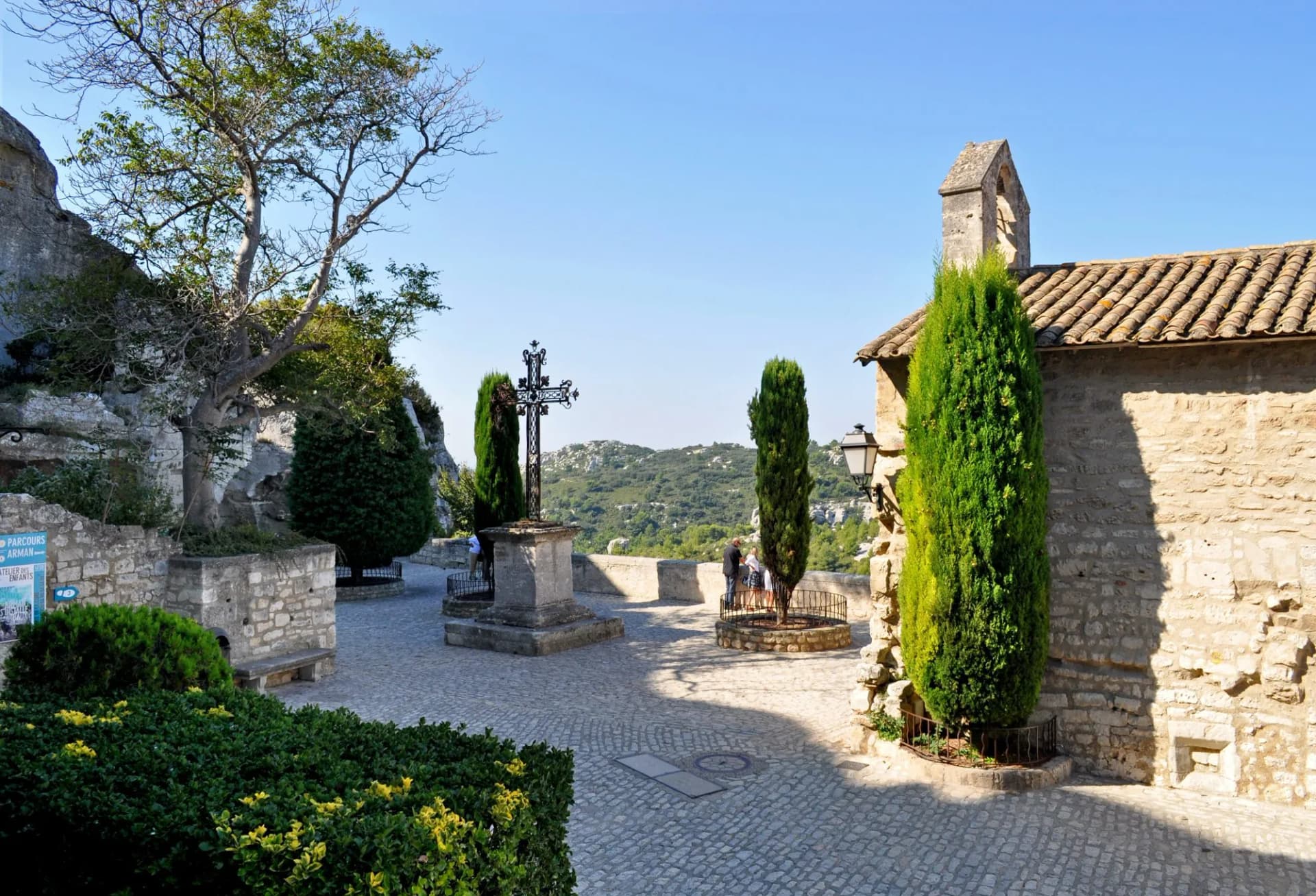 Cobblestone square with stone cross, cypress trees, and stone building in Les Baux-de-Provence.