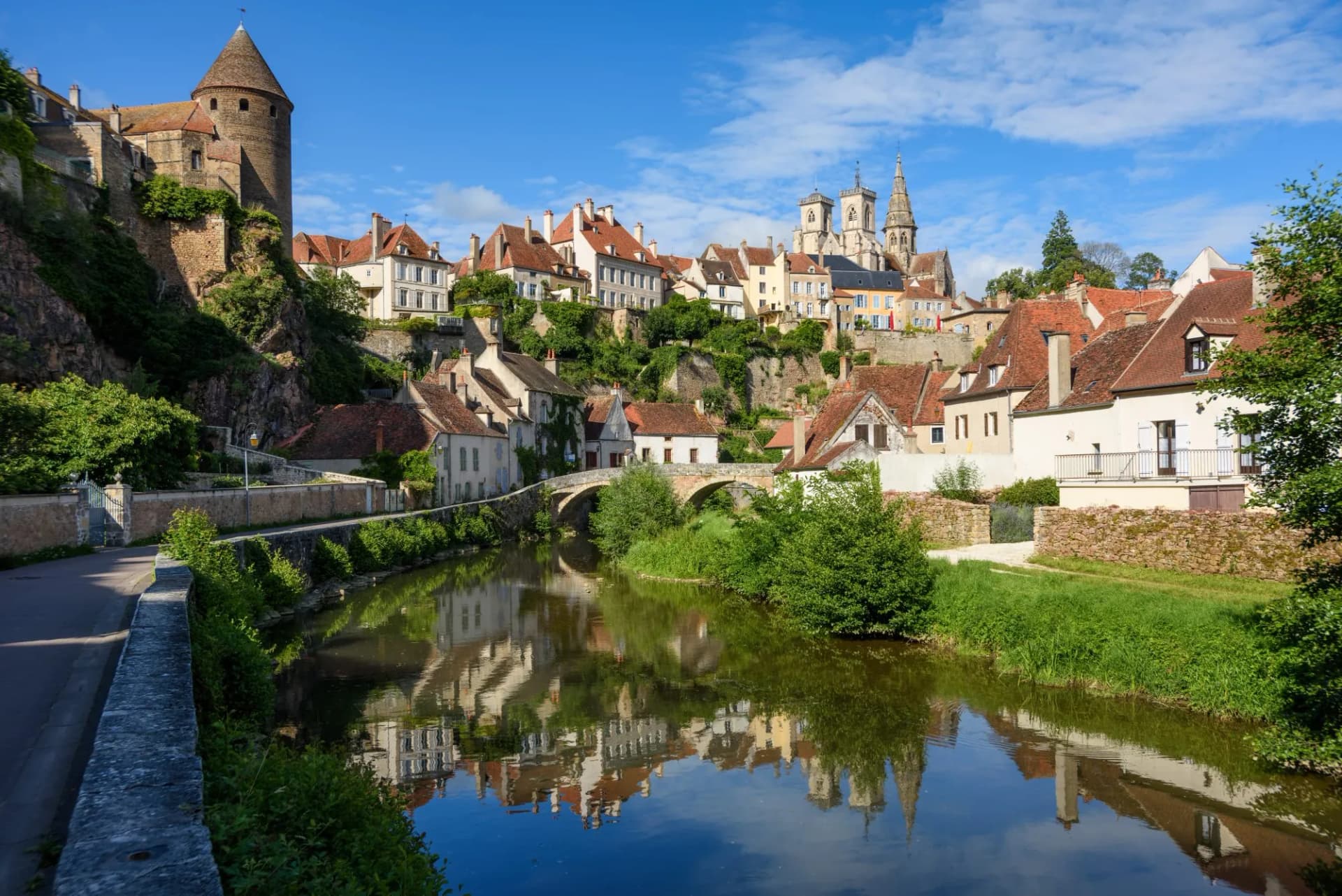Historic town with stone tower and church reflected in river under blue sky, Beaune.