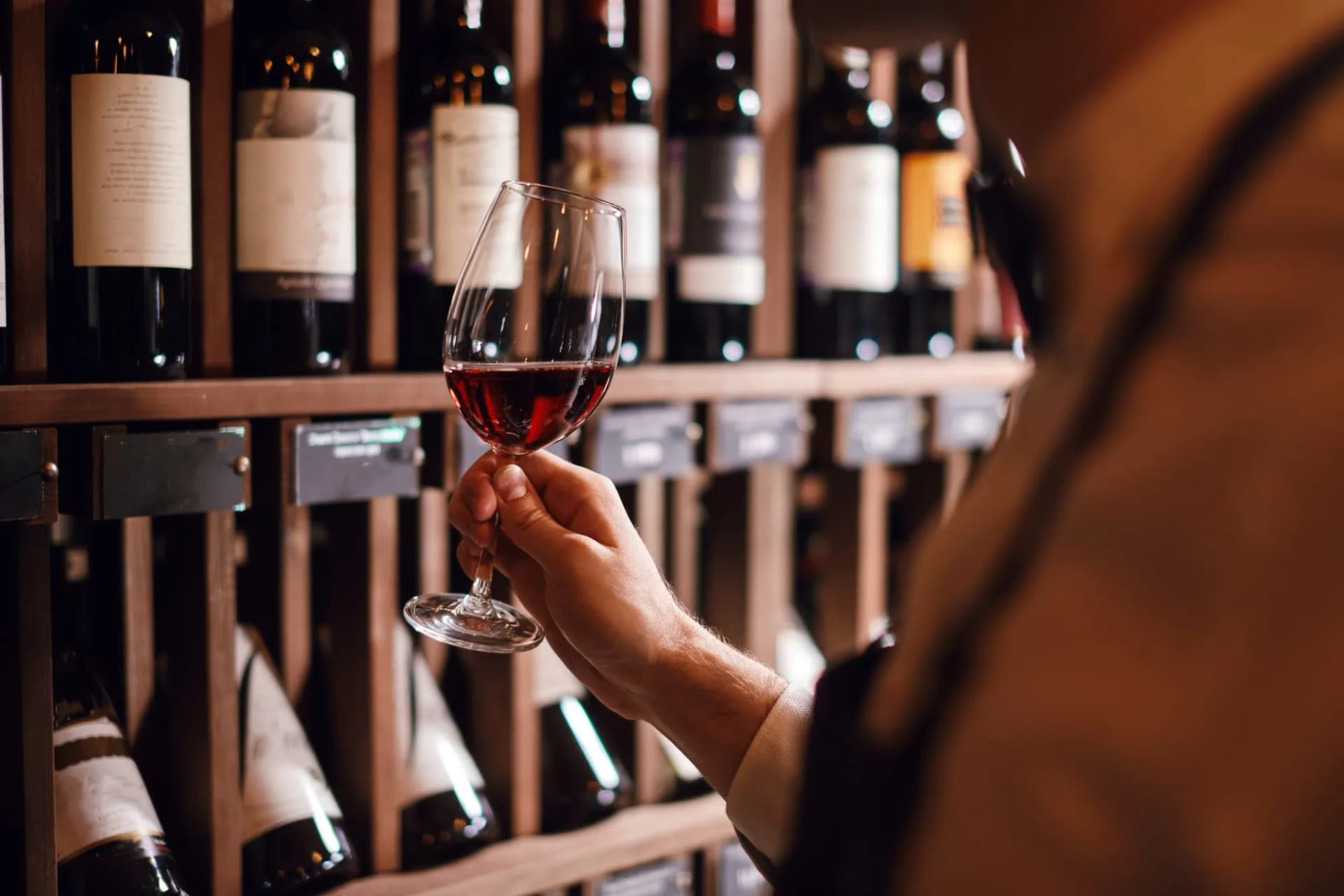 Hand holding glass of red wine while inspecting bottles on wooden wine rack in cellar