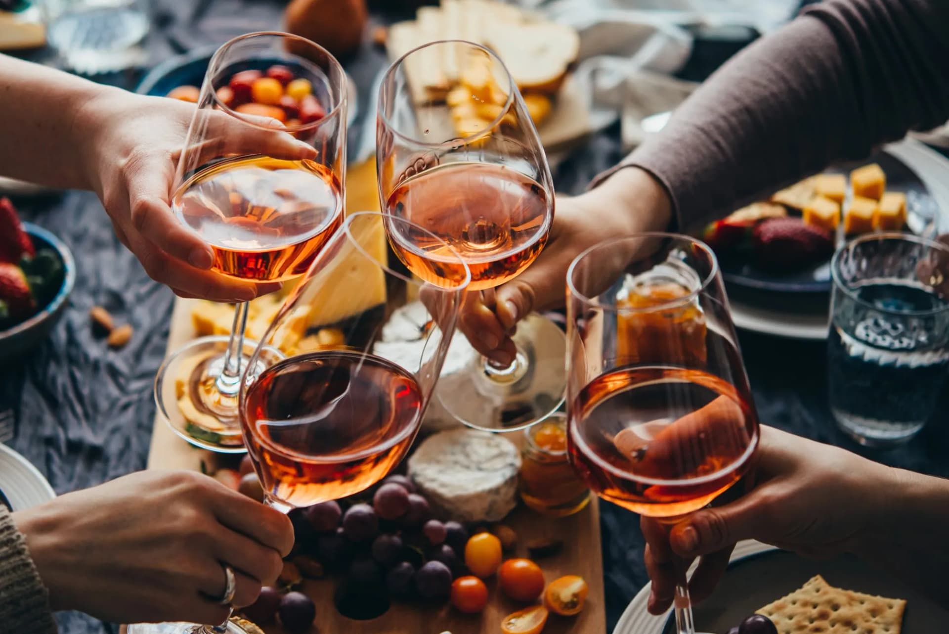 Hands toasting with rosé wine glasses over a cheese and fruit platter.