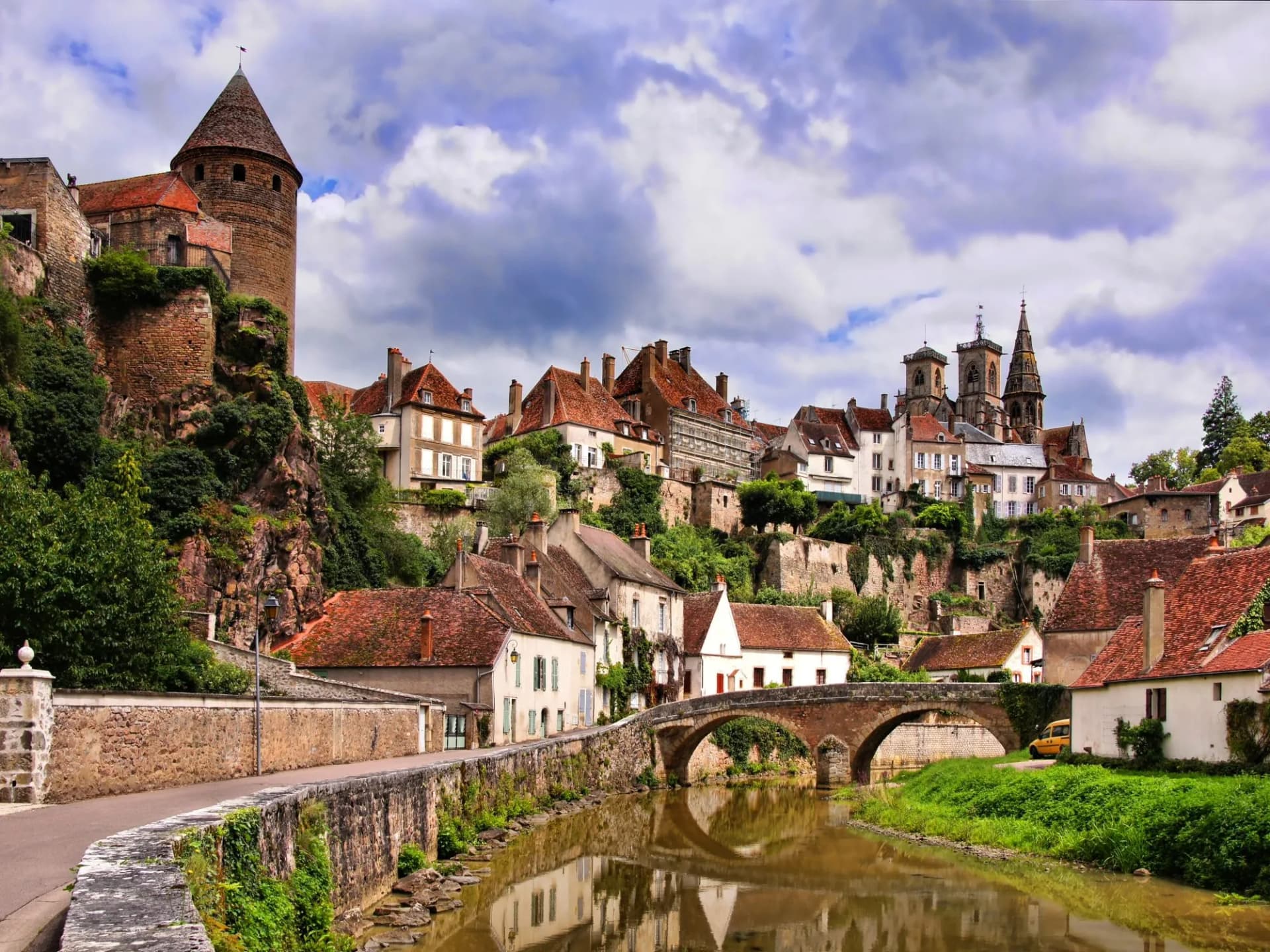 Historic town of Beaune with stone tower, bridge, and church overlooking a river under a cloudy sky.
