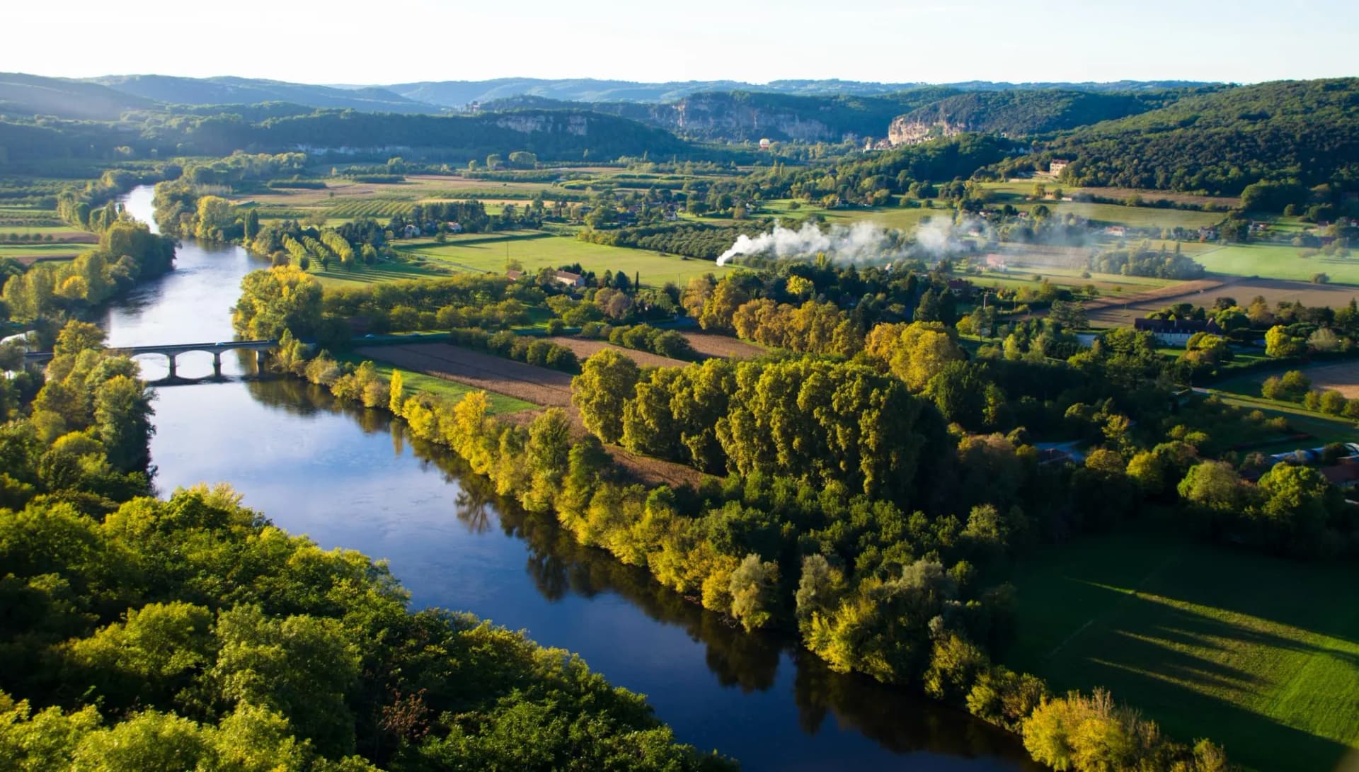 River winding through green valley with autumn trees, bridge, and distant cliffs in Dordogne.