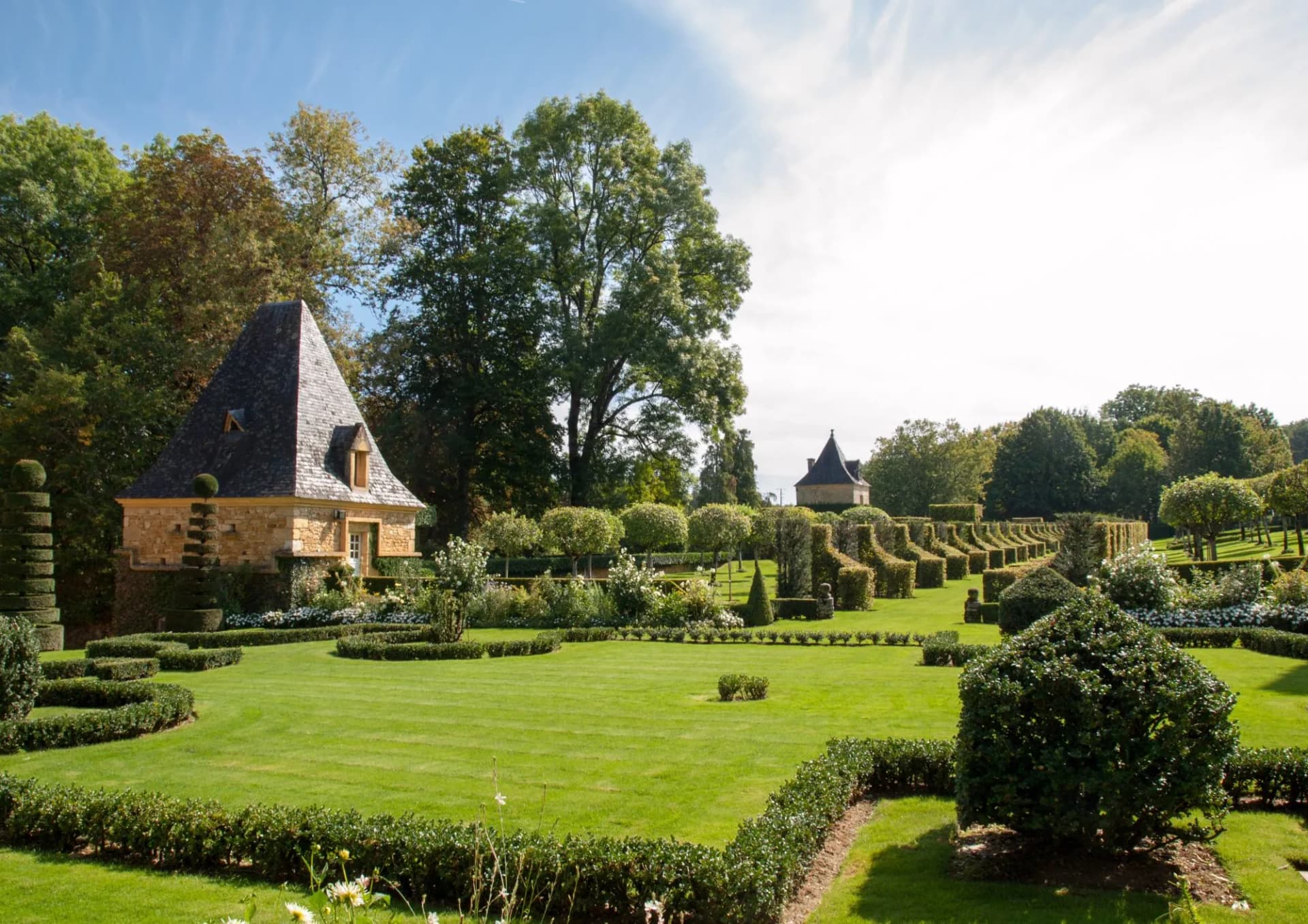 Formal garden with manicured hedges, topiary, and a stone building under a bright sky.
