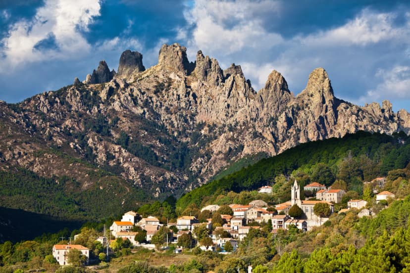 Village nestled in green hills below jagged rocky mountains under a blue cloudy sky.