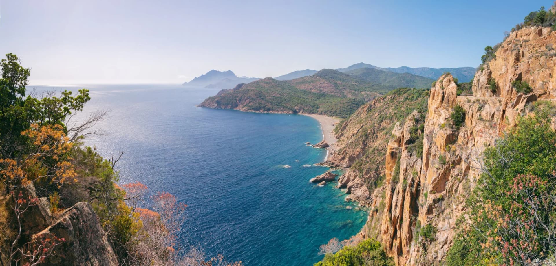 Coastal view of steep orange cliffs, blue sea, and distant mountains in Porto, Corsica, France.