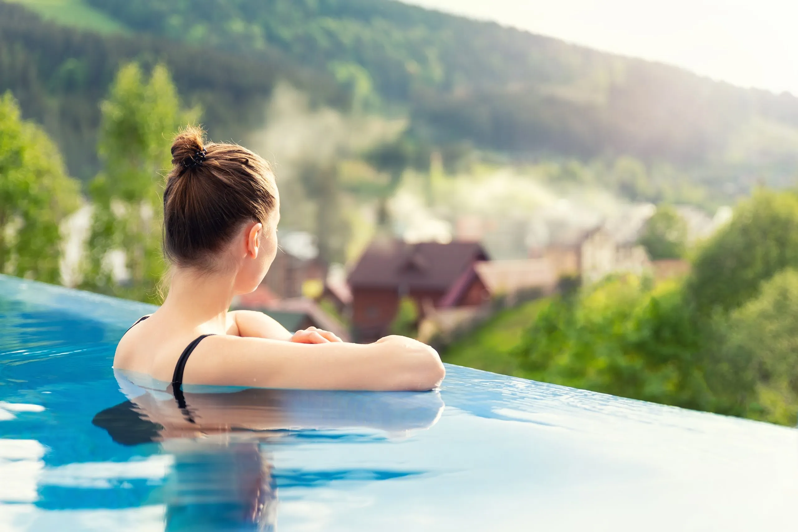 Back view young adult female person enjoy relax in infinity edge luxury outdoor swimming pool looking on fog hill green mountains in warm sunset light. Alpine welness travel vacation tourism concept.