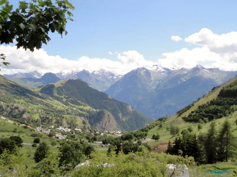 Blick von Alpe d'Huez, Frankreich, an einem sonnigen Sommertag. Die Vegetation auf dem Berg ist üppig und grün, und der Himmel ist strahlend blau mit flauschigen, weißen Wolken. In der Ferne sind die schneebedeckten Berge der Alpen zu sehen.